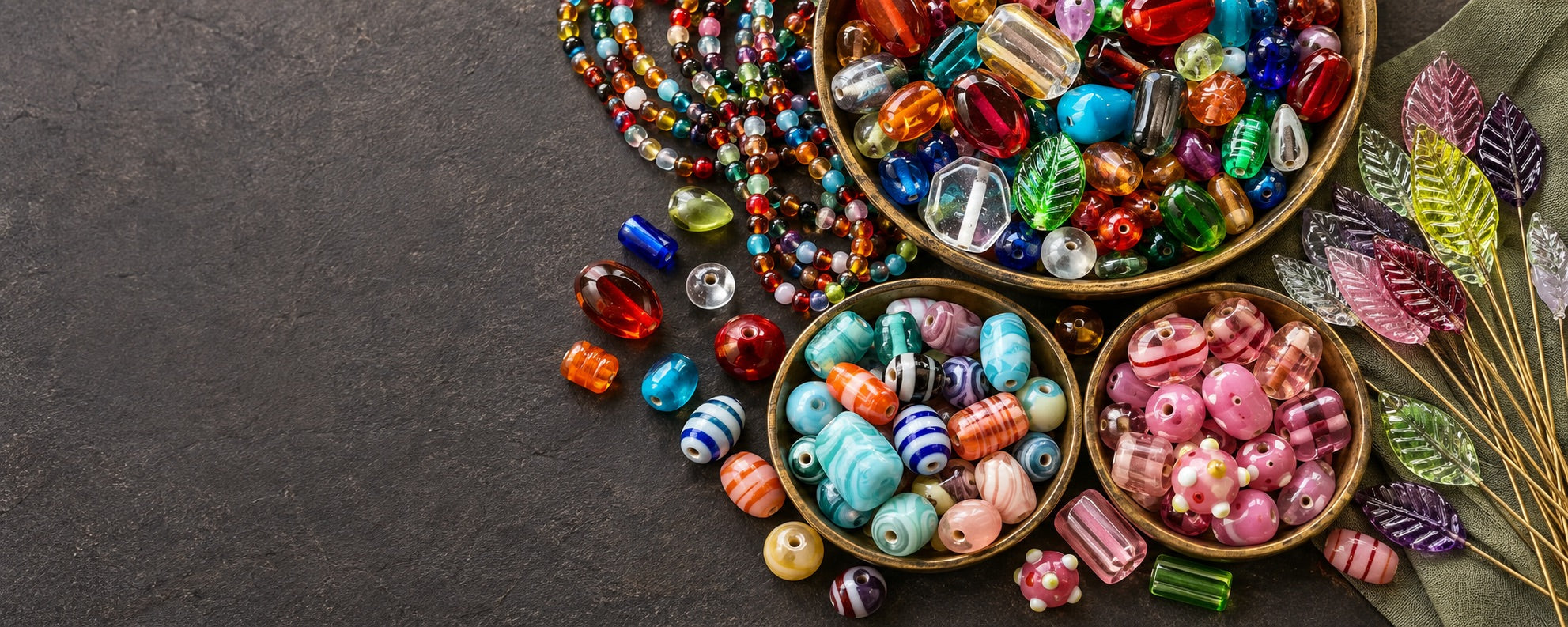 Bowls of colorful glass and lampwork beads, bead strands and glass leaf headpins on a dark surface